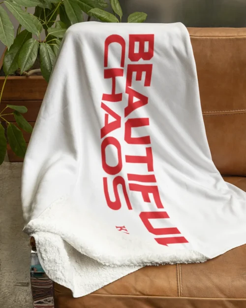 white katseye blanket with red beautiful chaos lettering laying on a brown couch